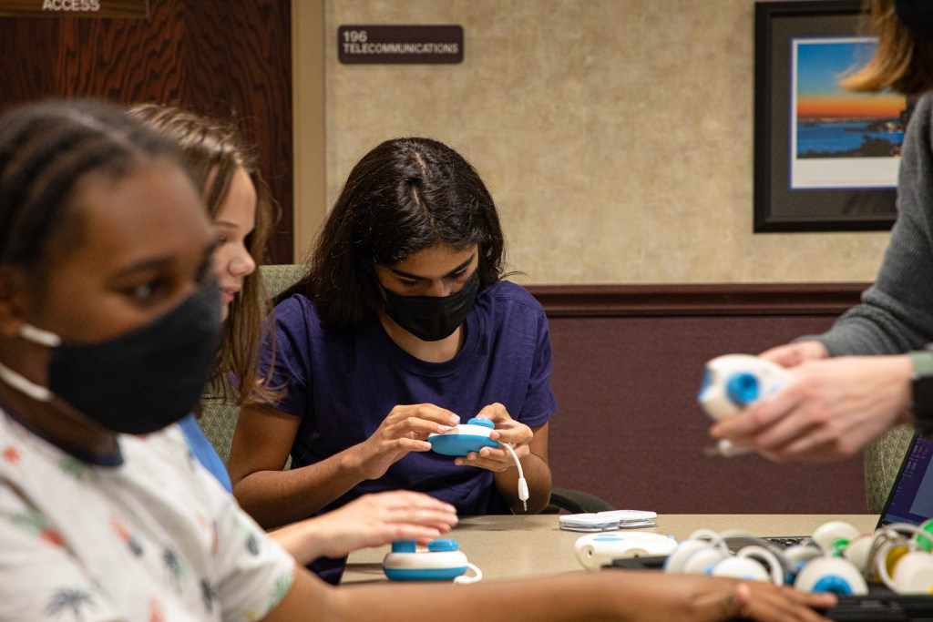 Fifteen-year-old Diego Zamora Blanco holds a small blue device that teaches students coding using music. Around him, two other students examine the same hardware with the help on an instructor.
