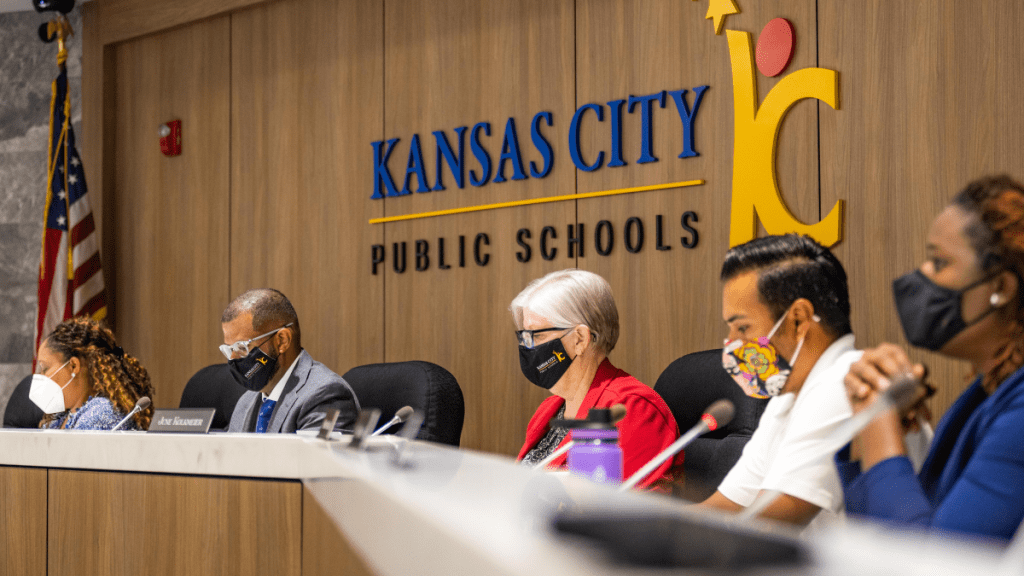 Image of five Kansas City School Board members seated at a desk during a board meeting. A large sign reading "Kansas City Public Schools" is visible behind them.
