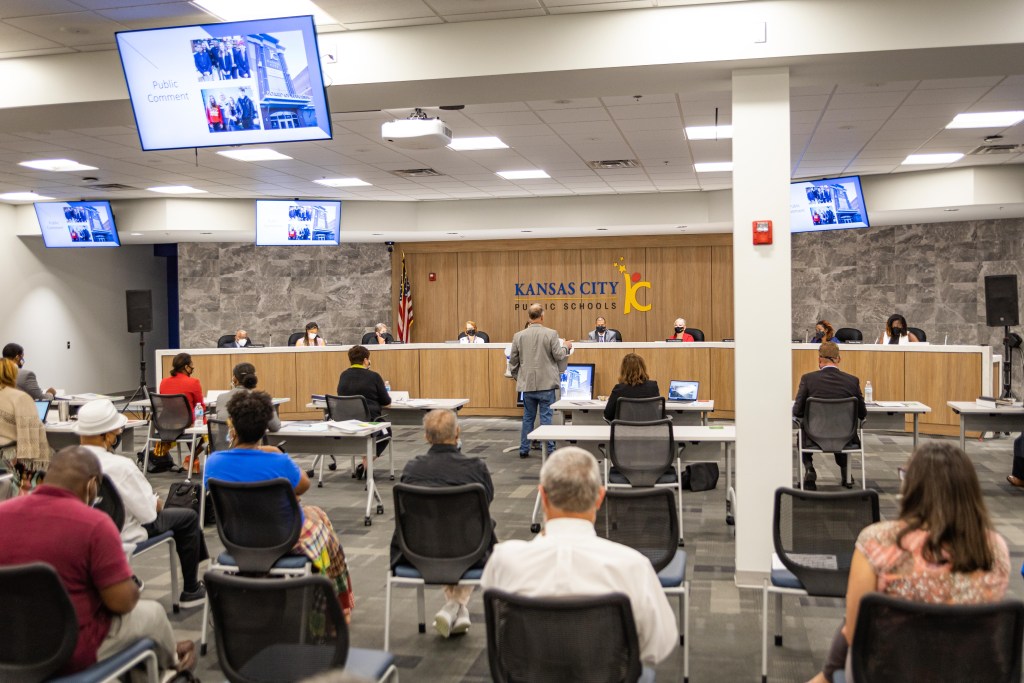 Image of the large room where Kansas City Public Schools board meetings are held. Board members are seated at a large desk and members of the public are seated in chairs spread throughout the room.