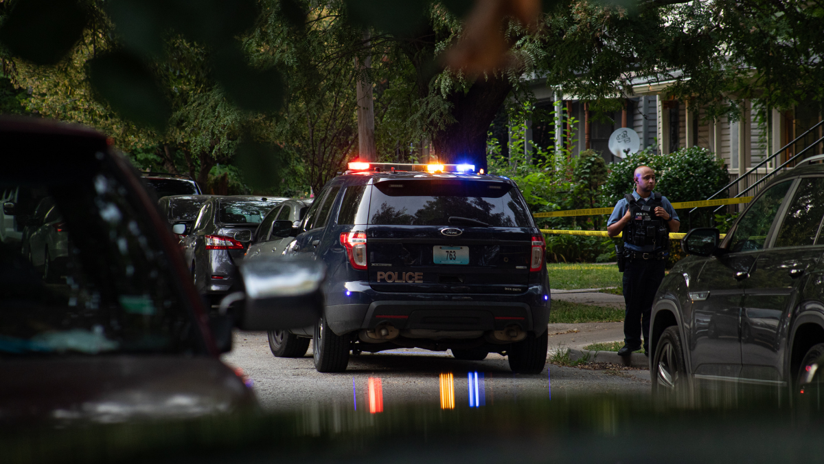 A police car with its lights on, pulled over on the side of a residential street. An officer stands outside on the sidewalk.