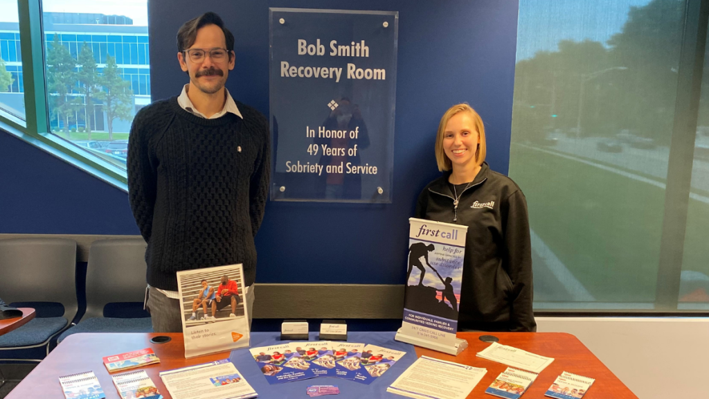 Two people stand in front of a sign that says, "Bob Smith Recovery Room," with a table set up with pamphlets and flyers.