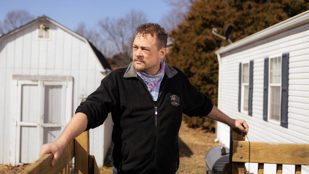 Robert Richardson, dressed in a jacket and tie-dye bandana, stands on the front porch of his trailer. His arms are holding onto the railing, and he's looking to his right.