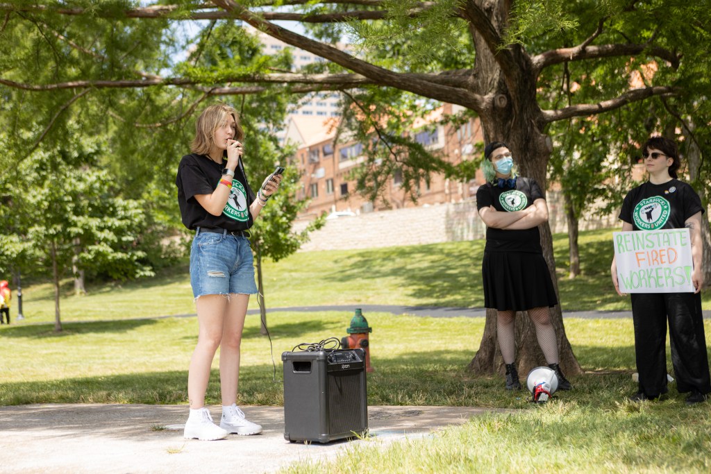 Three young people stand in a park wearing black Starbucks Workers United shirts. A woman speaks into a microphone next to a speaker.