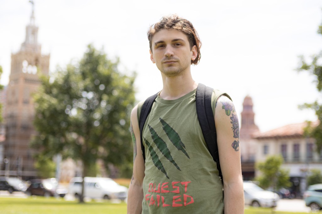 A young man in a green shirt wearing a backpack stands in a park near Kansas City's Country Club Plaza for a story about the Starbucks union effort.