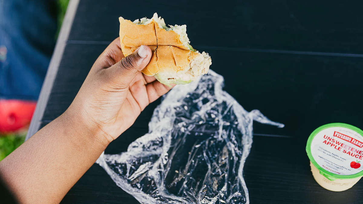 A child's hand hold's a half-eaten sandwich, with an unopened container of applesauce
