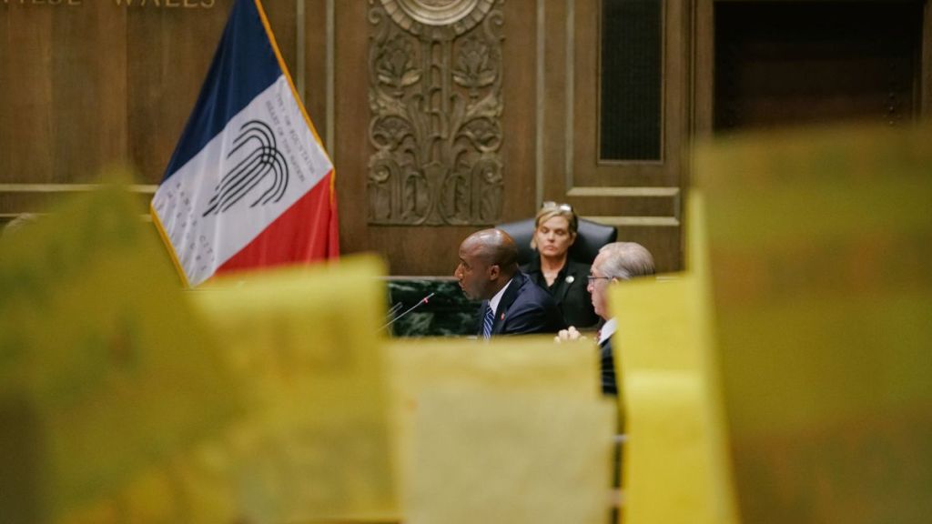 Mayor Lucas, seated, speaks into a microphone at City Hall. In the foreground, KC Tenants hold signs protesting the new housing ordinance.