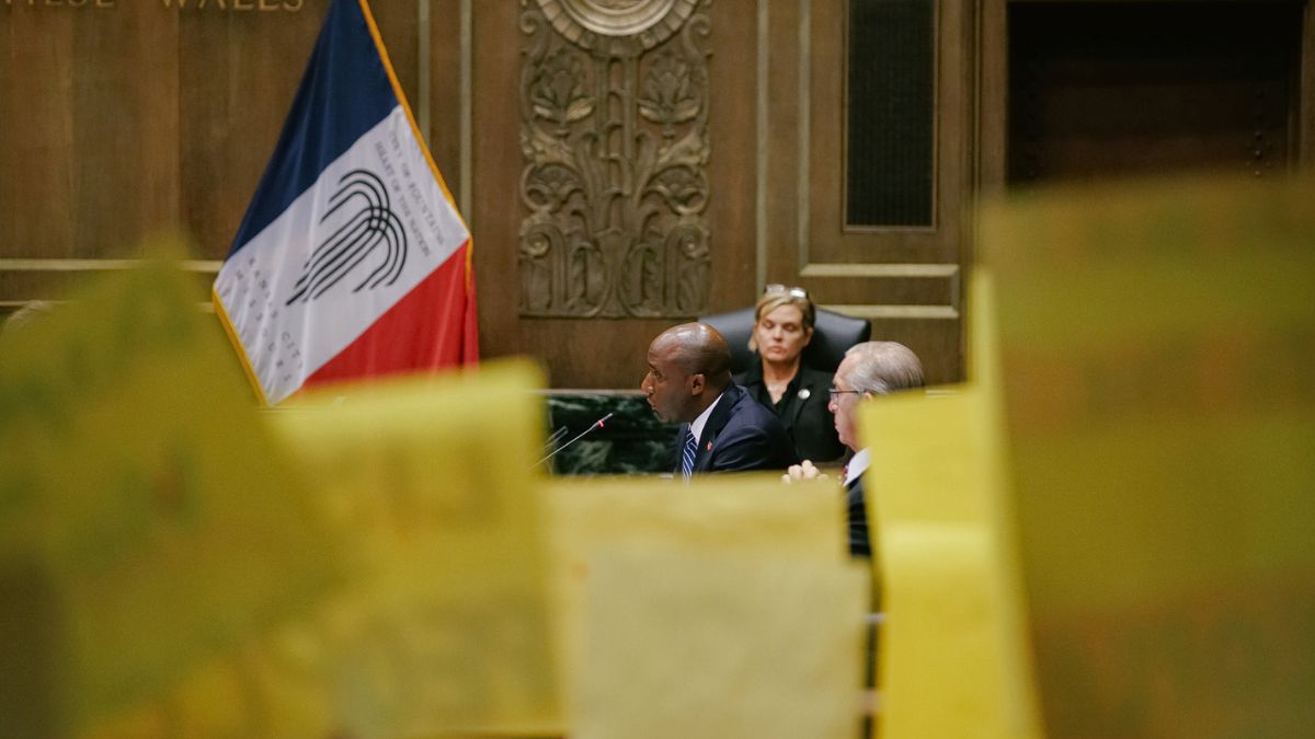 Mayor Lucas, seated, speaks into a microphone at City Hall. In the foreground, KC Tenants hold signs protesting the new housing ordinance.