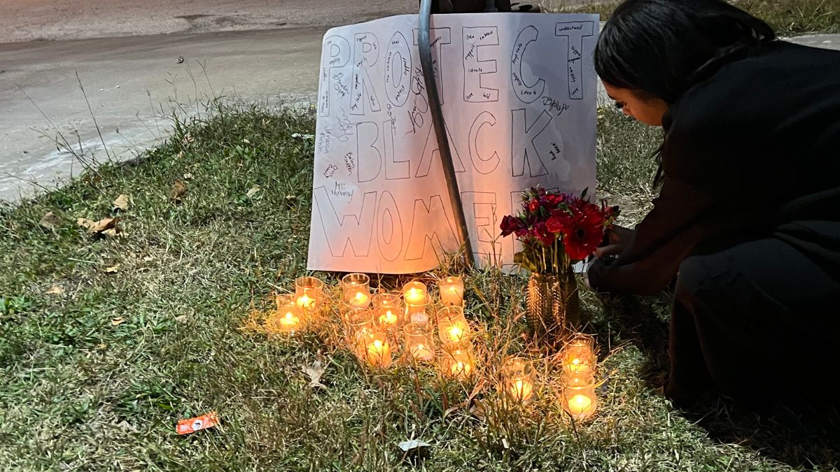 A woman kneels down beside a sign that reads "Protect Black Women." In front of the sign are a bunch of lit candles and a vase of roses.