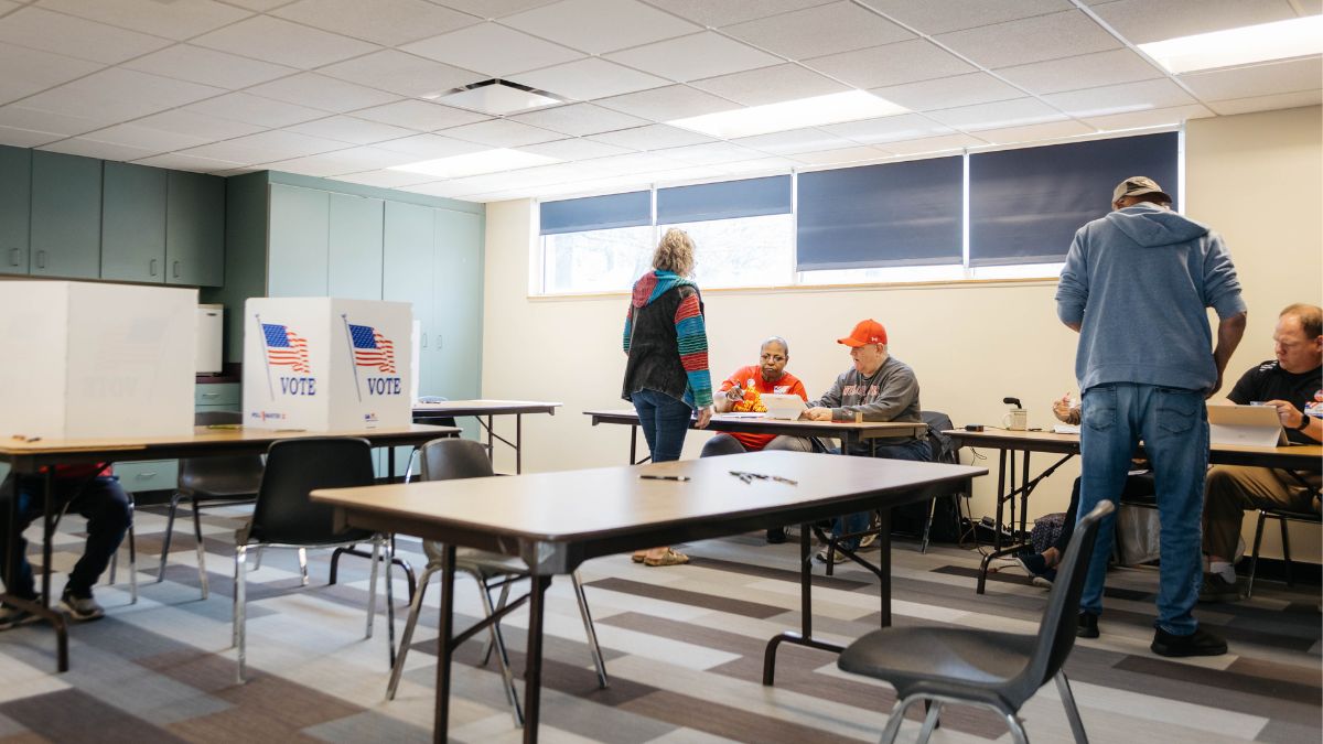 A polling location in Kansas City North Community Center, where four workers are pictured greeting voters, and one person fills out a ballot to the left.