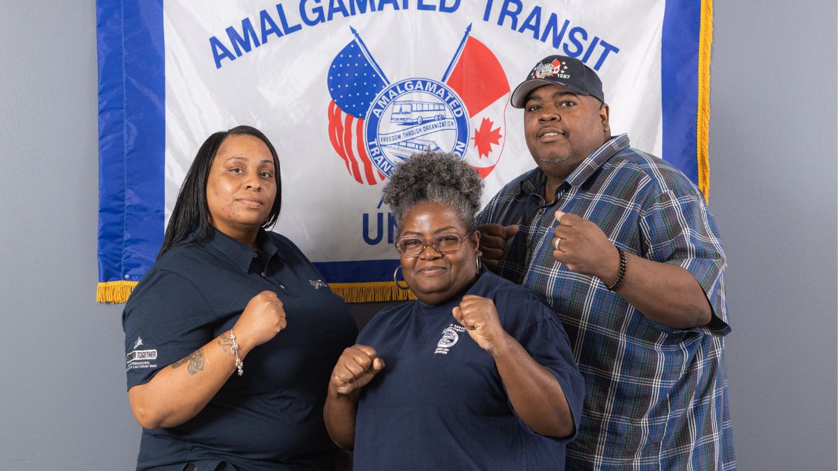 Three members of the Amalgamated Transit Union 1287 stand in front of their union flag, raising their fists.