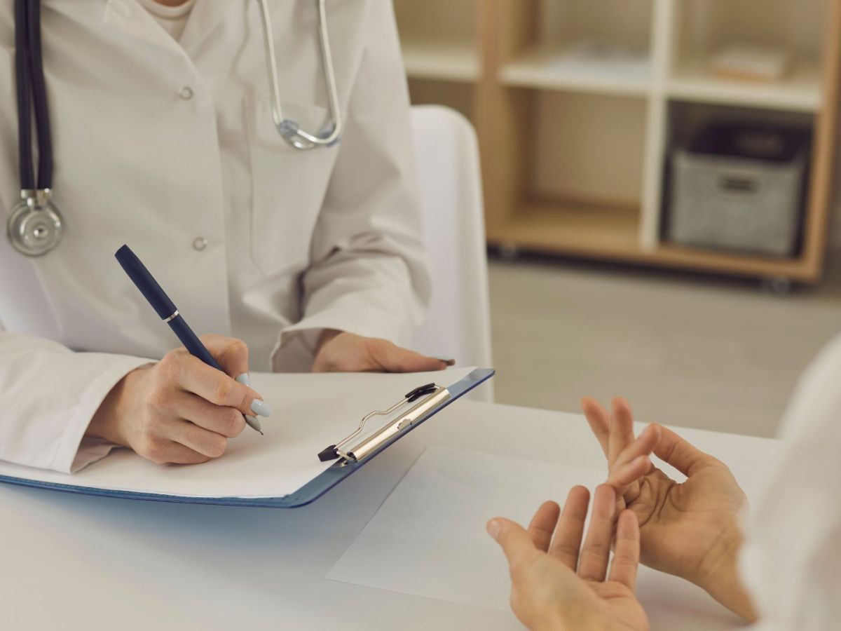 A medical professional in a white lab coat fills out a form as a person in the foreground gestures with their hands