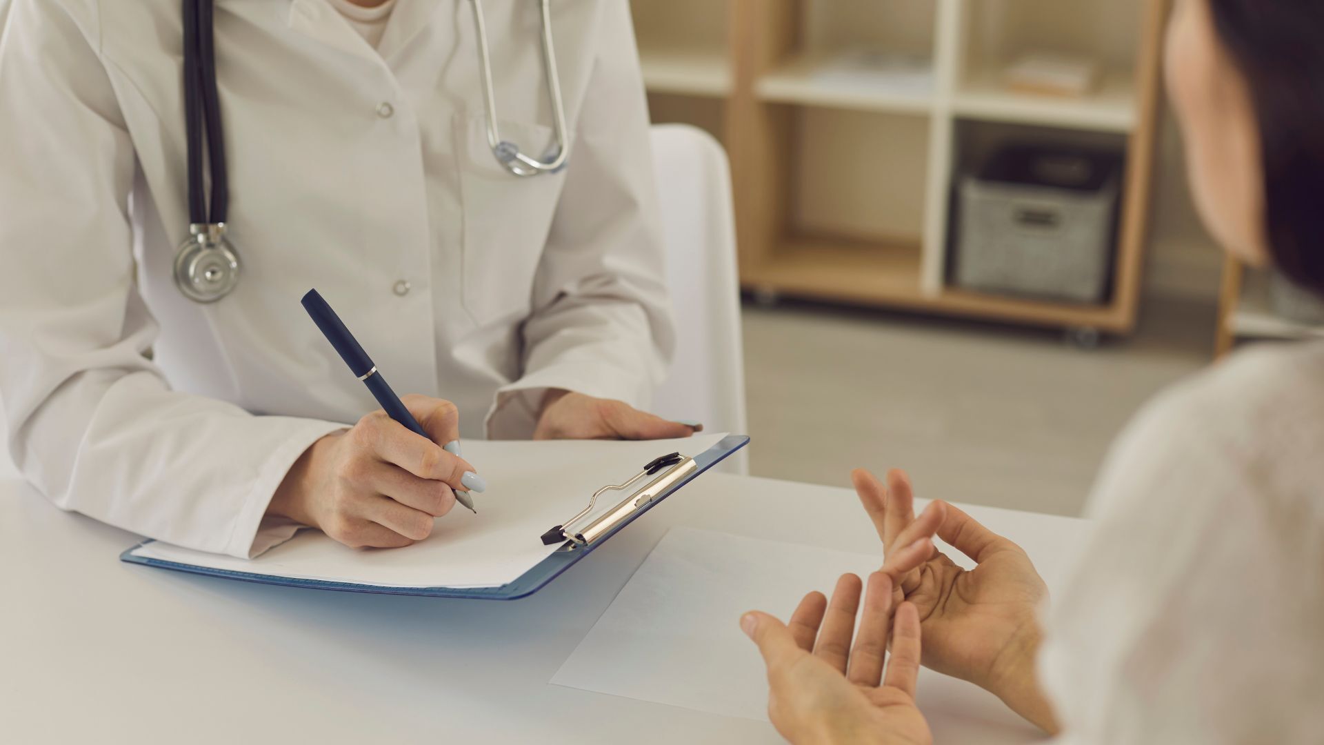 A medical professional in a white lab coat fills out a form as a person in the foreground gestures with their hands