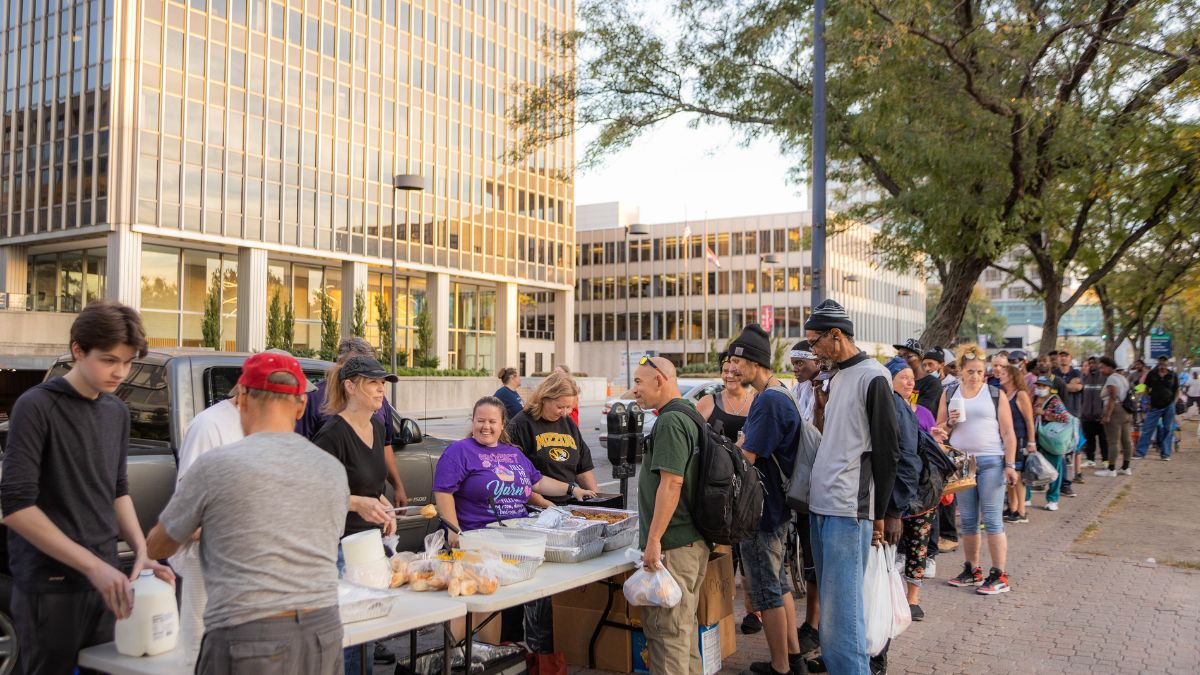 A group of volunteers serves food to a line of Kansas City unhoused people in the park.