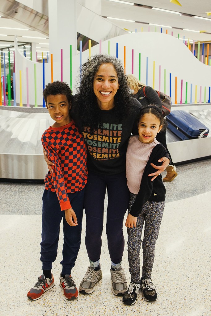 Robynn Nichols stands in the middle, with one kid on either side, in front of a baggage claim carousel decorated in bright colors.