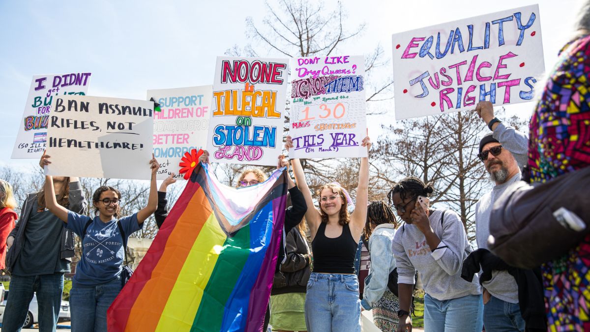 A group of protesters outside hold hand-made signs and a rainbow flag.