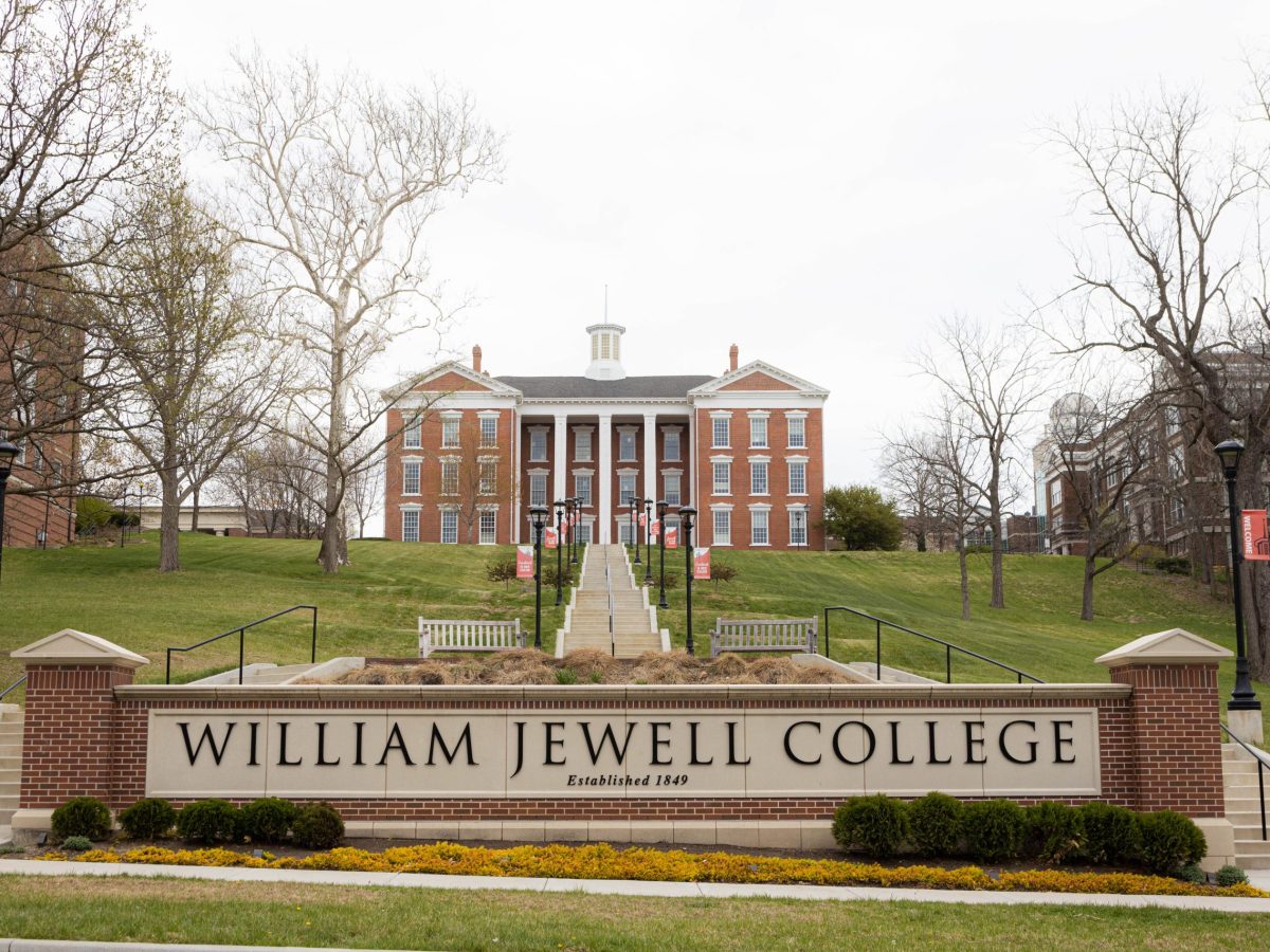 A sign reads William Jewell Collage in front of the exterior of Jewell Hall, a large brick building with columns surrounded by trees.
