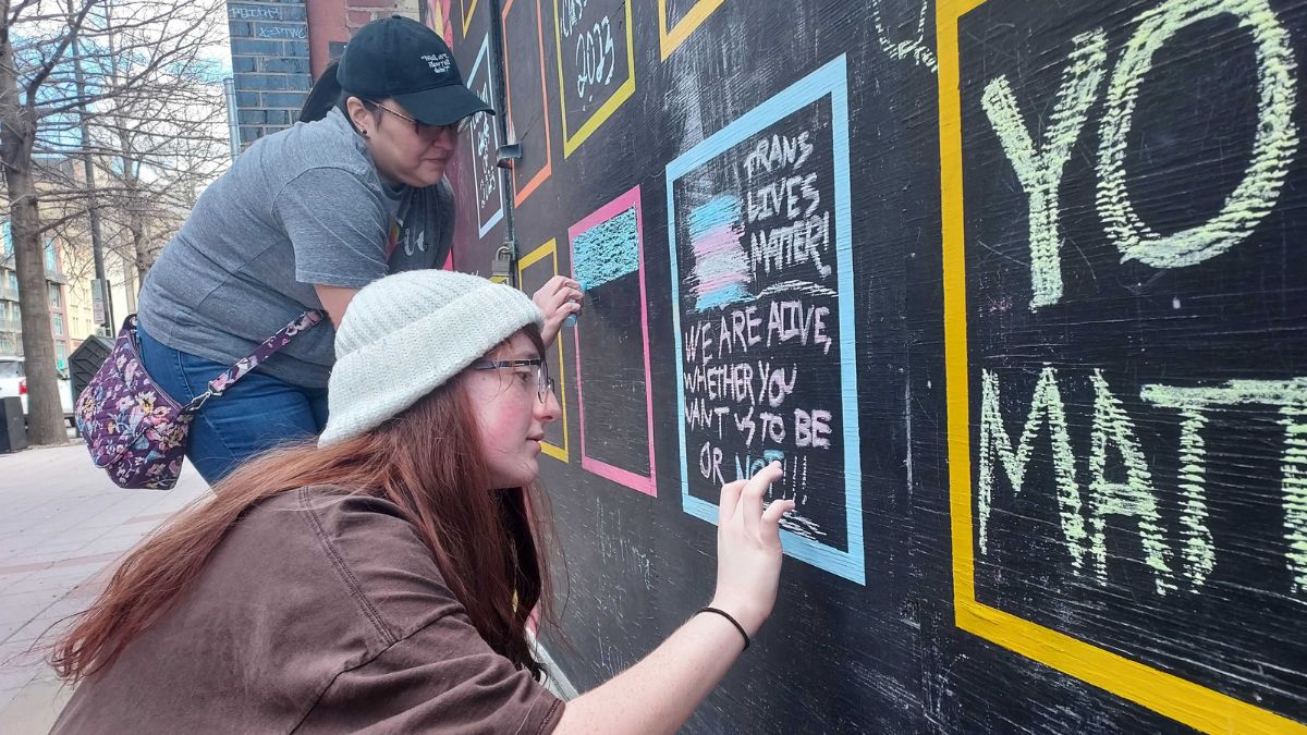 People draw chalk messages on a large black board outside.