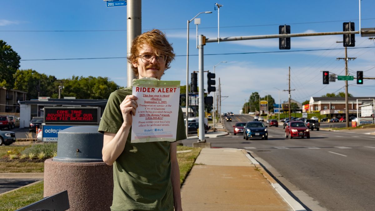 A portrait of Jaz Hays at a Gladstone bus stop. He is holding up a notice about bus service ending in the area.