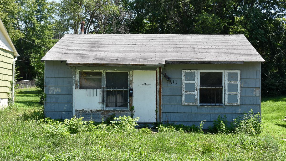 A grey house sits in a overgrown lawn with a "no tresspassing" sign on the door.