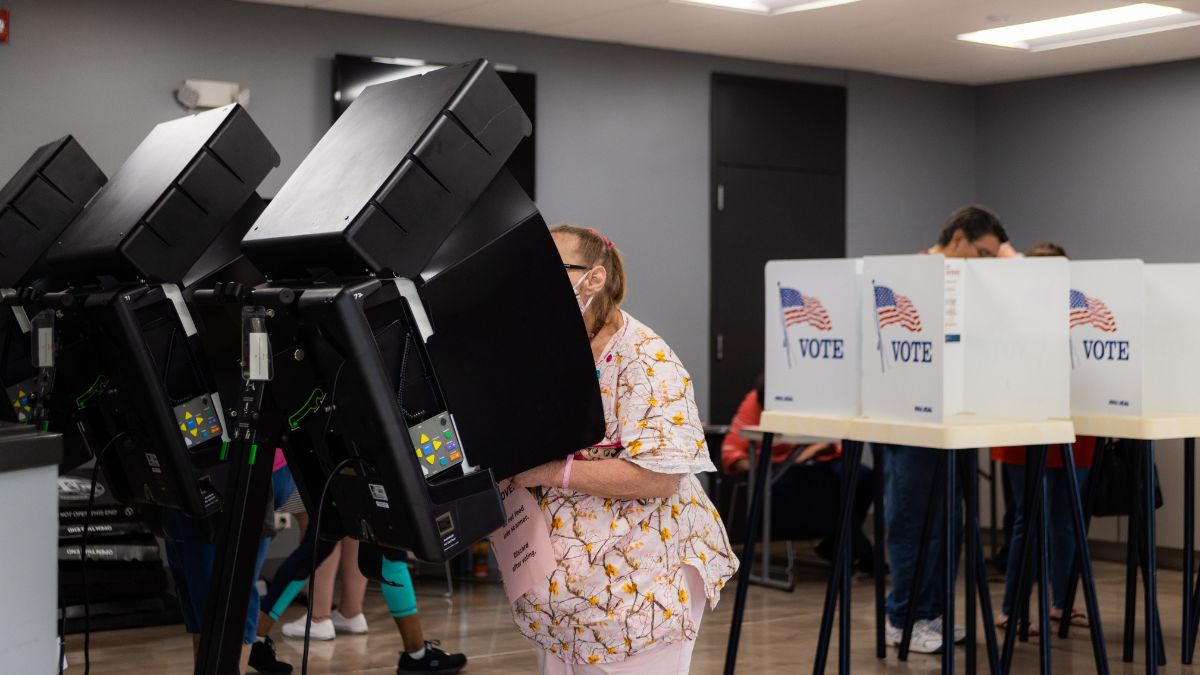 A voter operates a voting machine in the foreground. Other voters cast paper ballots at booths in the background.