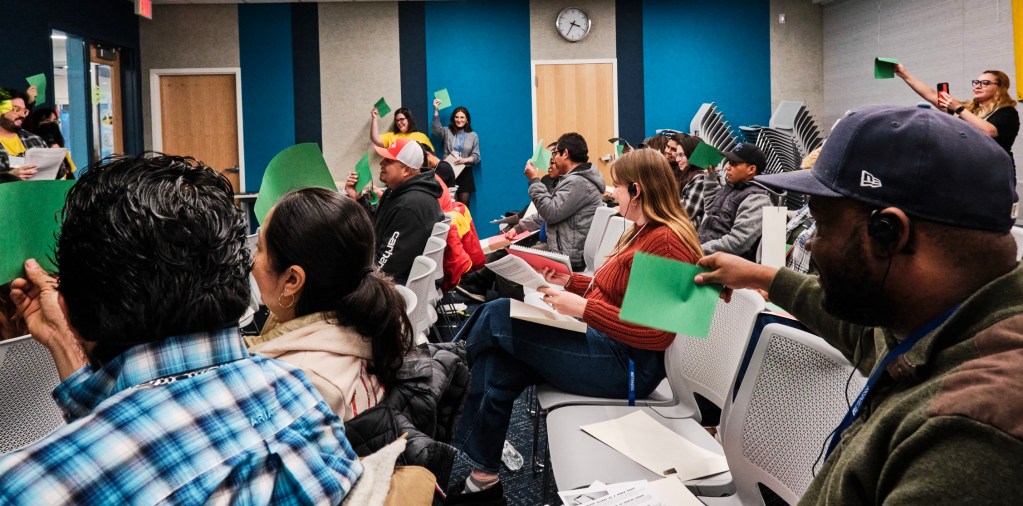 A crowd holds green papers up to answer yes-or-no questions from the speaker. Around two dozen people are visible, with some wearing earpieces for interpretation.
