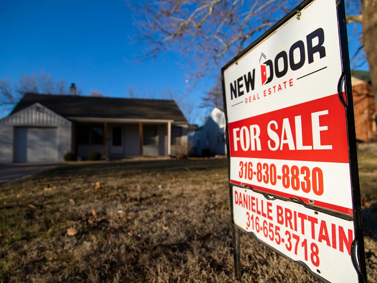 A red and white real estate sign in a yard in front of a gray one story house.