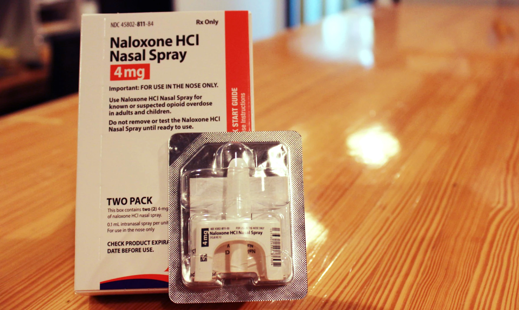a white box titled "Naloxone HCI Nasal Spray" sits on top on a table next to the spray itself.