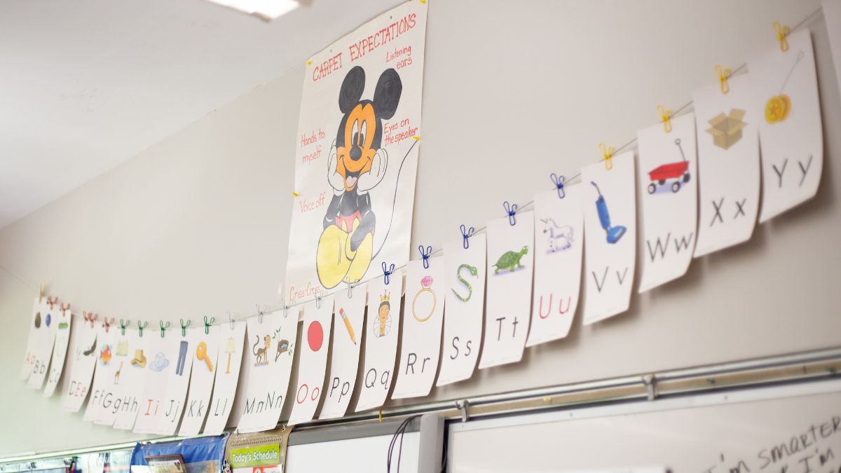 Classroom wall with alphabet cards, each showing a letter and matching image, strung on a clothesline. A poster with Mickey Mouse lists classroom behavior expectations like “Hands to myself” and “Voice off.”