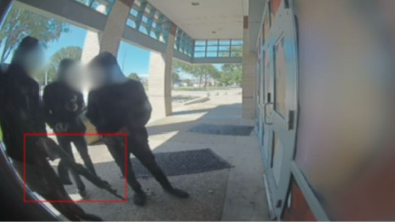 The photo shows four individuals standing outside a building entrance. Their faces are blurred, and they are dressed in dark clothing, with one person holding an AK-47, highlighted by a red box.