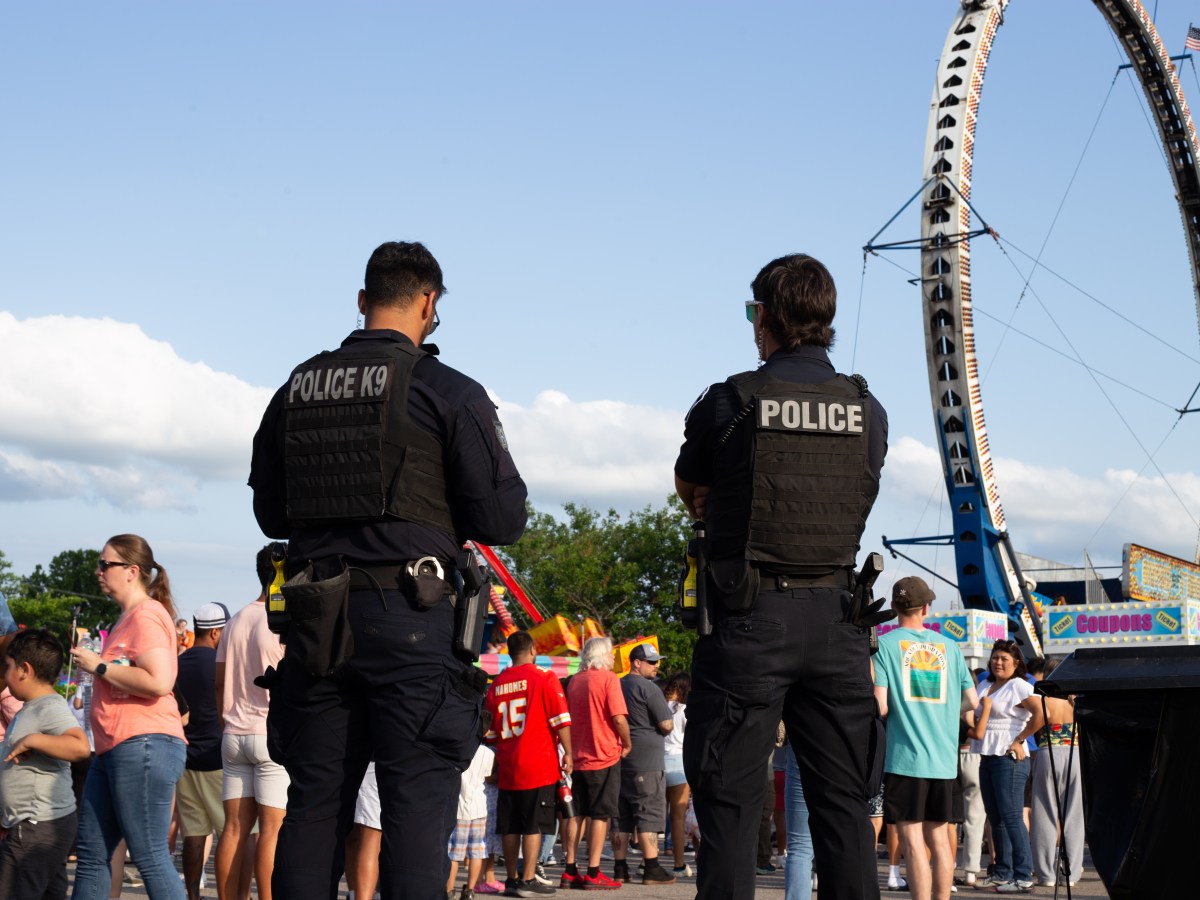 In this photo, two police officers wearing tactical vests stand with their backs to the camera, overseeing a crowded outdoor event. In the background, there are numerous people walking around, a tall circular amusement park ride and booths.
