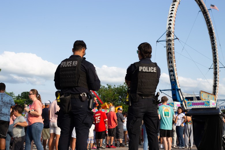In this photo, two police officers wearing tactical vests stand with their backs to the camera, overseeing a crowded outdoor event. In the background, there are numerous people walking around, a tall circular amusement park ride and booths.