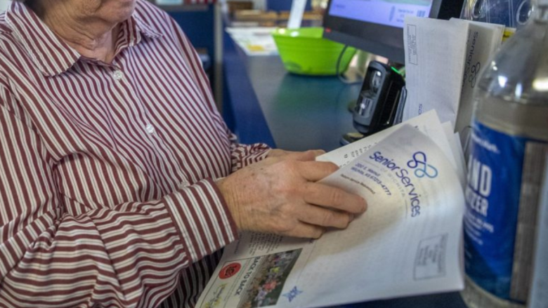 An older person wearing a red and white striped shirt is standing at a counter, holding a stack of papers. One paper on top says “Senior Services” and has a phone number printed on it. Nearby, there is a bottle of hand sanitizer and a green bowl on the counter next to a card reader.