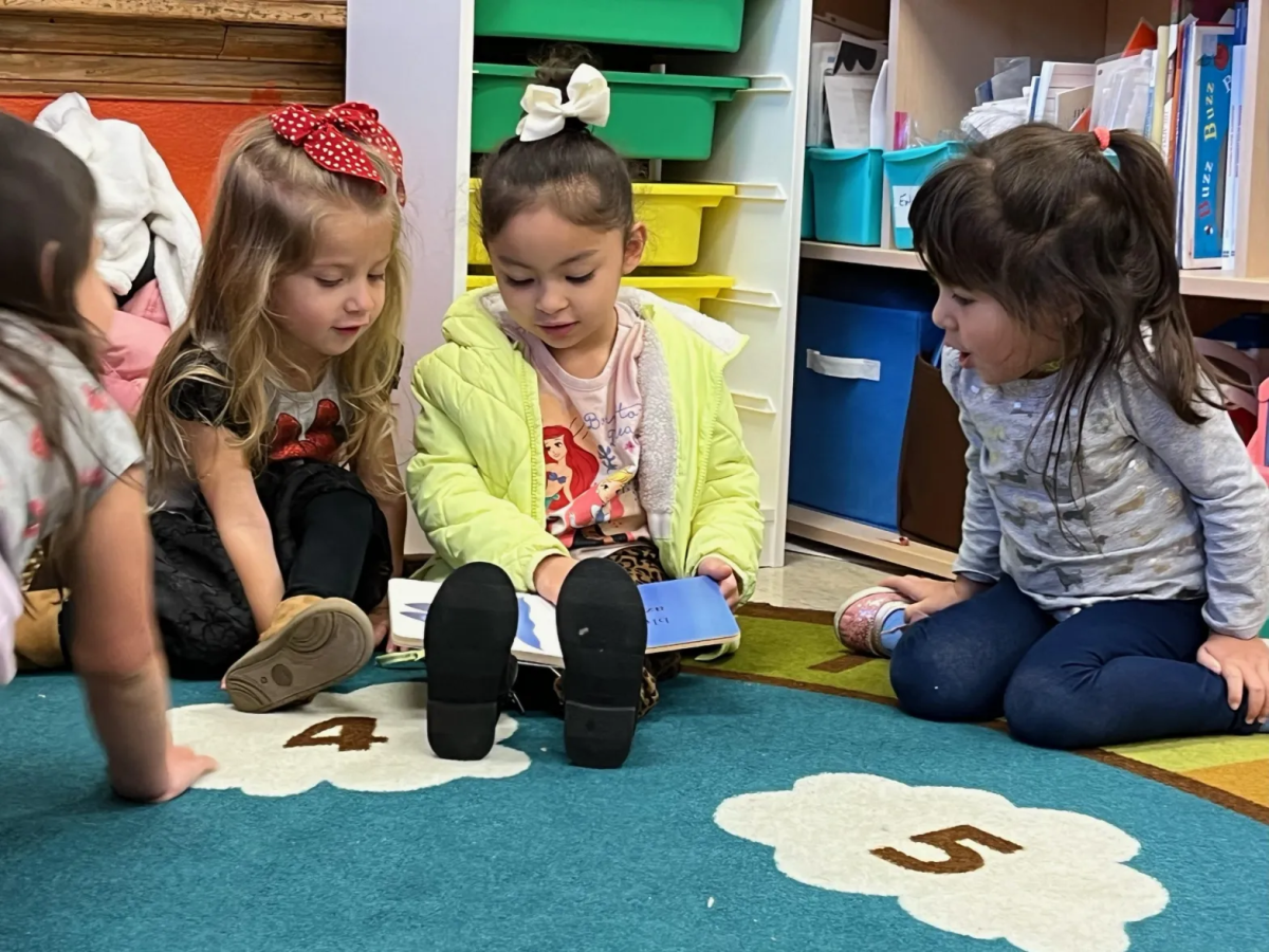 Four young girls are sitting on a classroom rug, gathered closely as one of them in a light green jacket reads aloud from a book held open in her lap. The girls are surrounded by colorful storage bins and bookshelves.