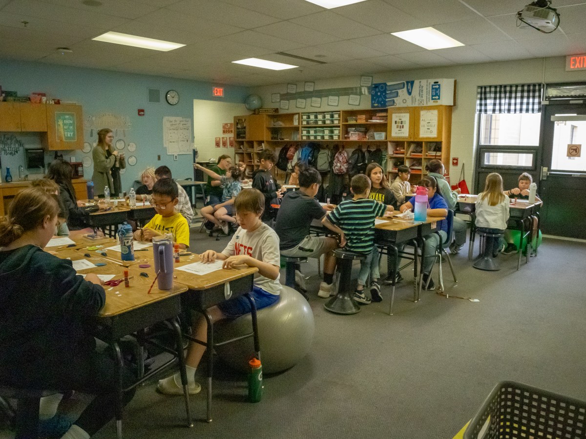 This photo shows a classroom with many young students sitting in groups at their desks, working on assignments or activities. A teacher stands at the front of the room while the students appear engaged and focused, some chatting or smiling.