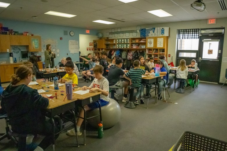 This photo shows a classroom with many young students sitting in groups at their desks, working on assignments or activities. A teacher stands at the front of the room while the students appear engaged and focused, some chatting or smiling.