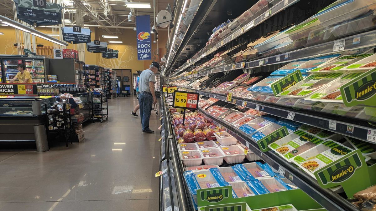 This photo shows the meat section of a grocery store, where neatly stacked packages of various meats—including ground turkey from Jennie-O—fill several shelves along a long refrigerated aisle. A man in a gray shirt and cap is closely inspecting items, and another person in yellow is working behind a display of discounted goods. Overhead signs indicate aisle numbers and product categories, while a University of Kansas "Rock Chalk Jayhawk" banner adds a local touch.