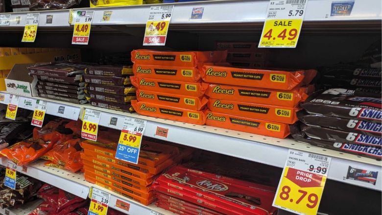 a grocery store candy aisle with various chocolate candy multi-packs on shelves. Brands like Reese’s Peanut Butter Cups, Hershey’s, Snickers, and Kit Kat are visible, with bright sale tags showing discounted prices.