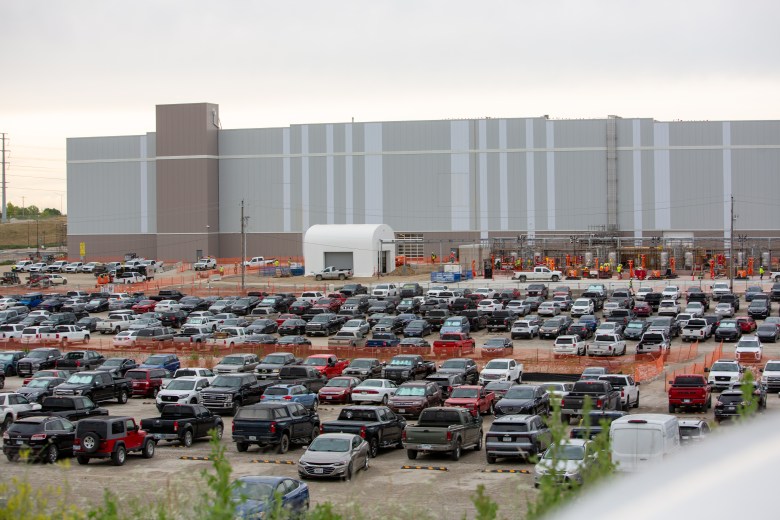 This photo shows the large, partially constructed De Soto facility with dozens of workers and heavy equipment near the building's perimeter. In the foreground, hundreds of vehicles — mostly pickup trucks and SUVs — are parked in a dirt lot.