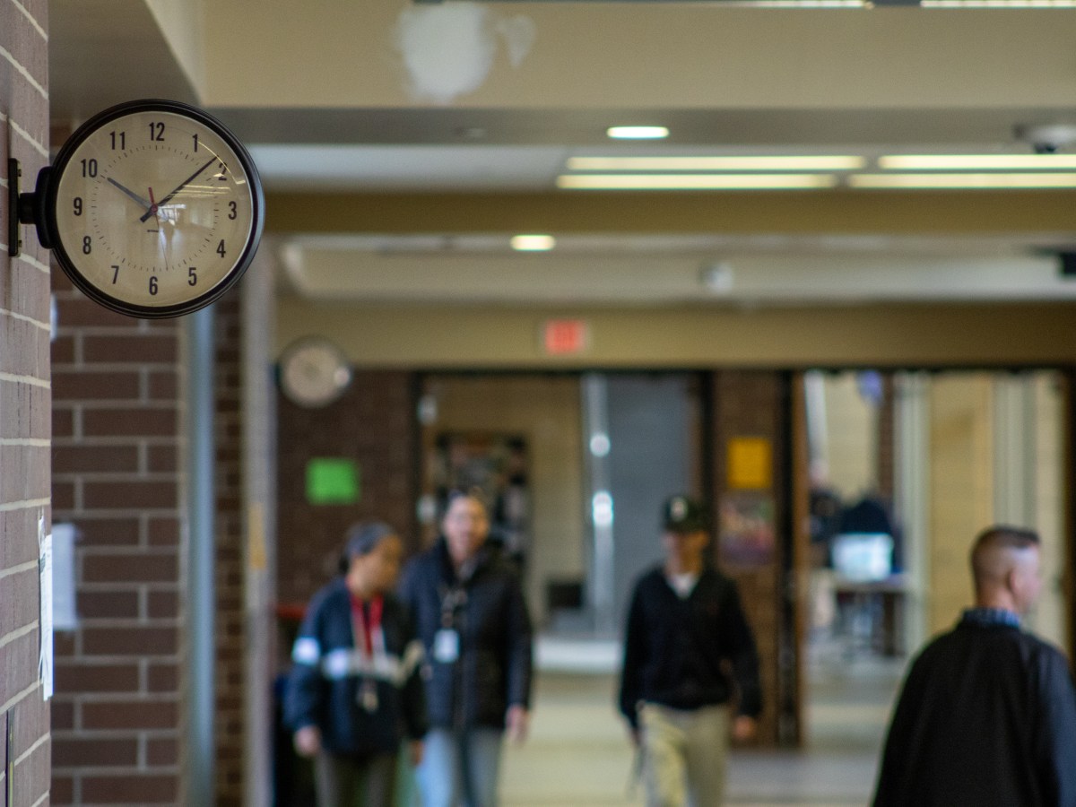 People walking through a school hallway. A clock sits in the upper lefthand corner.