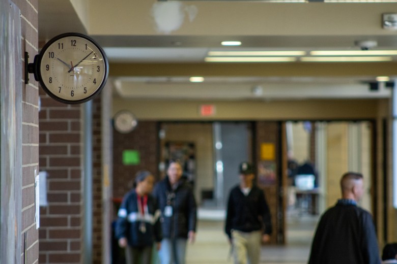 People walking through a school hallway. A clock sits in the upper lefthand corner.