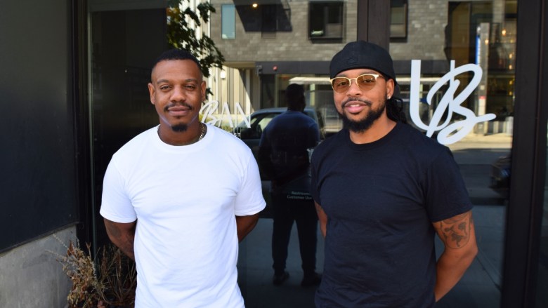 Two Black men pose together outside a modern storefront with large windows. The man on the left wears a white t-shirt, while the man on the right wears a black t-shirt, hat, and sunglasses. Reflected behind them is a contemporary building with multiple stories and balconies.