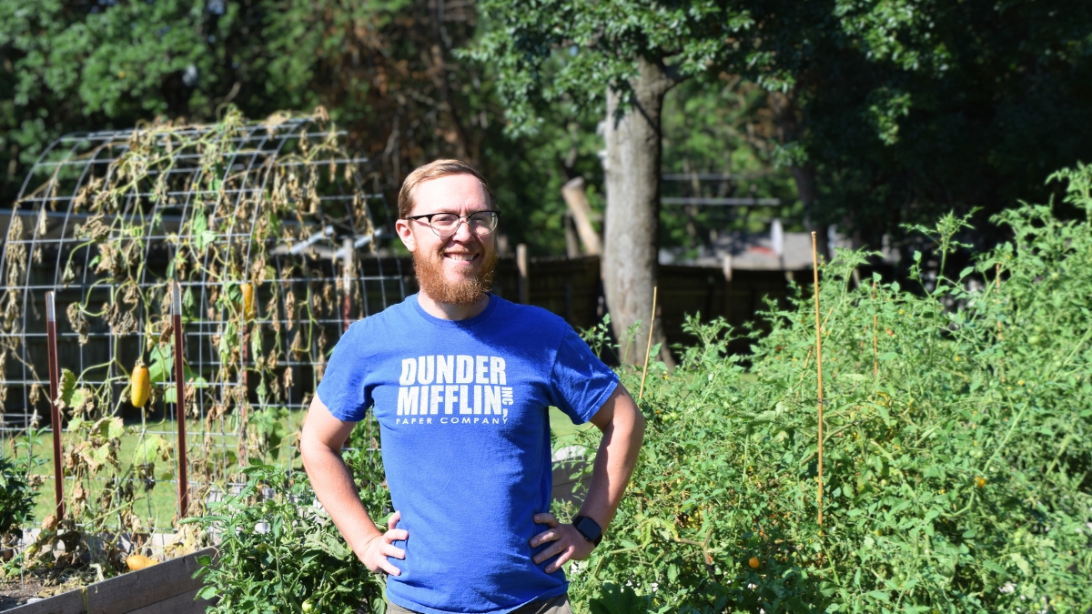 A smiling man with glasses and a red beard stands in a lush garden setting, wearing a blue t-shirt with 'DUNDER MIFFLIN PAPER COMPANY' text. Behind him are raised garden beds with various plants and vegetables, including what appears to be a trellis structure with climbing plants. The background shows mature trees and greenery