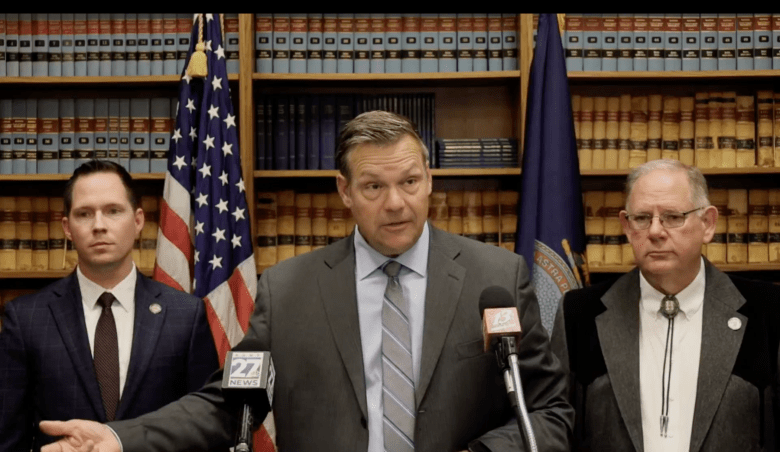 Kanas Attorney General Kris Kobach stands in front of a bookshelf. Dozens of books sit on the shelf, with the U.S. flag and Kansas flag next to him. Senate Majority Leader Chase Blasi and House Speaker Dan Hawkins are to his left and right.