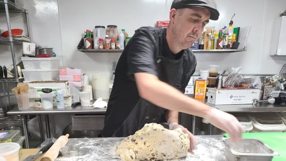 chef in black uniform and cap kneading dough on a floured work surface in a professional stainless steel kitchen with shelves of ingredients and equipment visible in the background.