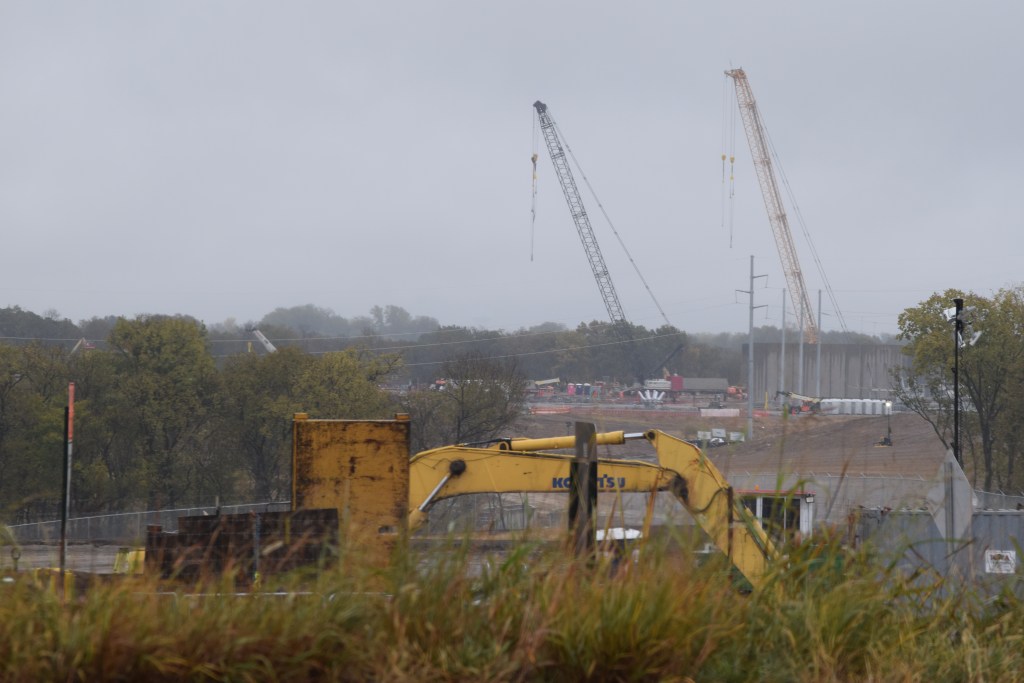 A yellow excavator sits beyond tall grass in the foreground with active construction visible in the background, including two large cranes and partially completed concrete structures under an overcast sky. Trees line the middle distance of the construction site.