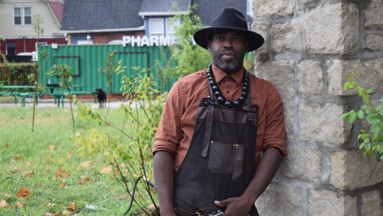 A Black man (Phillip Ramsey) wearing a black wide-brimmed hat, rust-colored button-down shirt, and black overalls with beaded necklace stands in a community garden with young fruit trees, green grass, and urban structures including a green shipping container and stone building in the background.