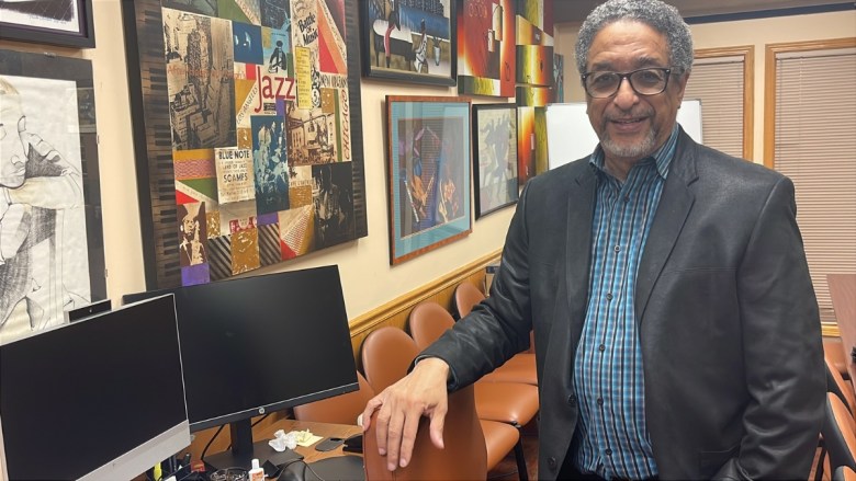 Clyde McQueen, a Black man with gray hair and glasses wearing a dark blazer and blue plaid shirt, leans on his chair while speaking in his office. Behind him are jazz-themed art posters and framed works on the wall, with dual computer monitors on a wooden desk.