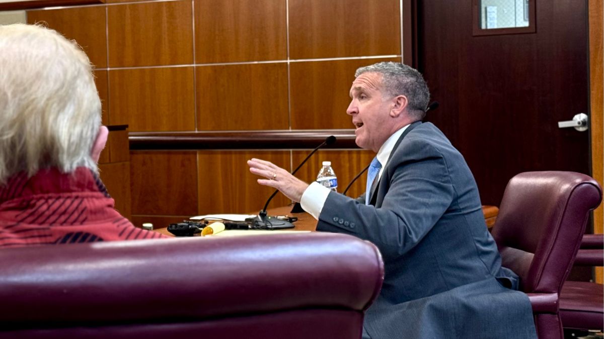 Phil LeVota, seated at a table in front of the county Legislature, speaks into a microphone, gesturing with his hands.