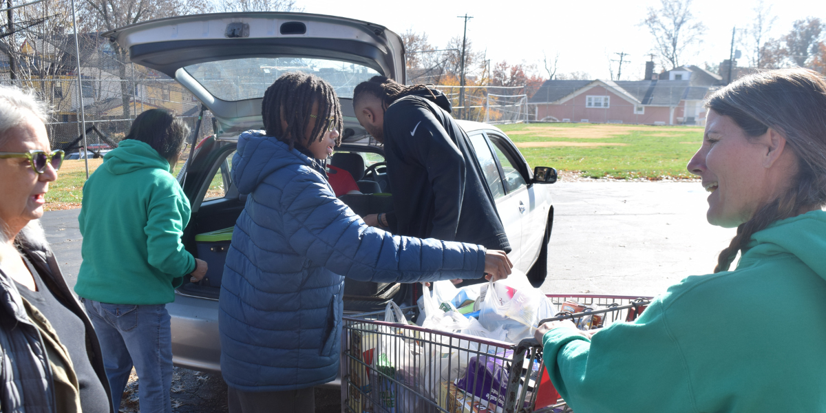 Volunteers hand food items from a car trunk to people with shopping carts at an outdoor food distribution event in Kansas City