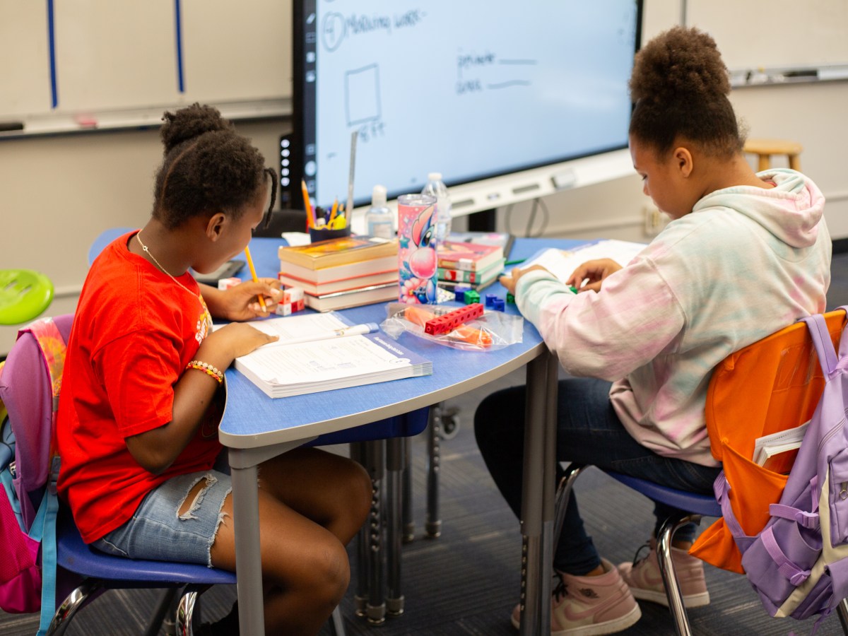 Students sit at a table in a classroom, writing in work books.